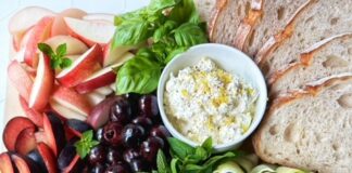 Cutting board with sliced tomatoes, zucchini ribbons, salami, nectarines, plums, peaches, pesto, buratta and ricotta cheese.