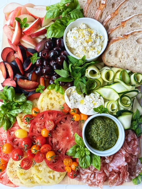 Cutting board with sliced tomatoes, zucchini ribbons, salami, nectarines, plums, peaches, pesto, buratta and ricotta cheese.