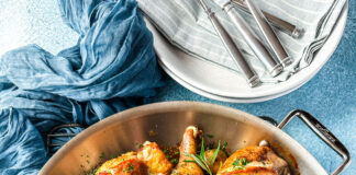 A saute pan with roasted chicken and grapes and a stack of plates and flatware.