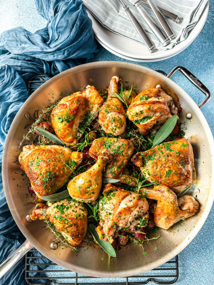 A saute pan with roasted chicken and grapes and a stack of plates and flatware.