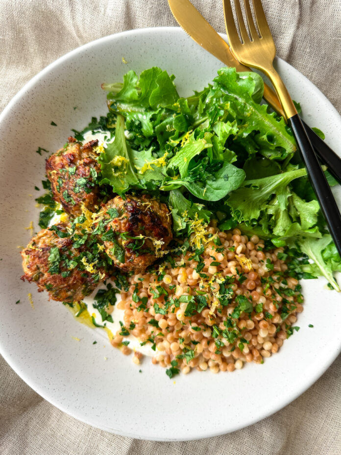 A shallow bowl of yogurt sauce topped with pearl couscous, meatballs and fresh greens.