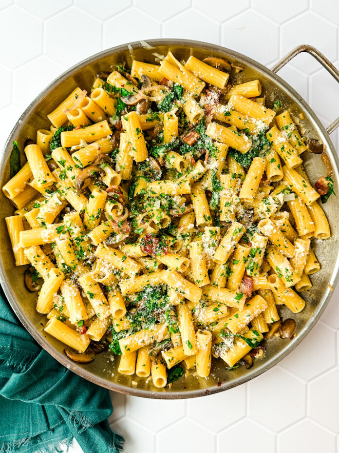 A saute pan filled with rigatoni pasta and bacon, mushrooms and spinach.