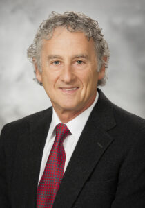 Dr. Neal Weinberg is a man with gray curly hair wearing a dark suit, white shirt, and red tie.