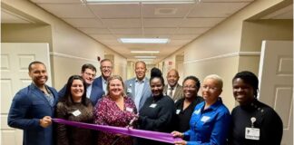 Group of racially diverse people in a hallway with an African American woman in a black dress holding the ceremonial "grand opening" scissors, cutting a purple ribbon.