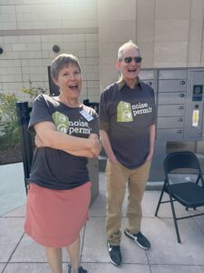 A man and woman wearing Noise Permit tee-shirts stand outside and smile to the camera