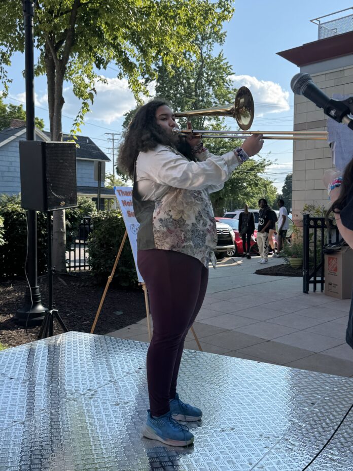 A young woman stands on an outside stage playing the trombone